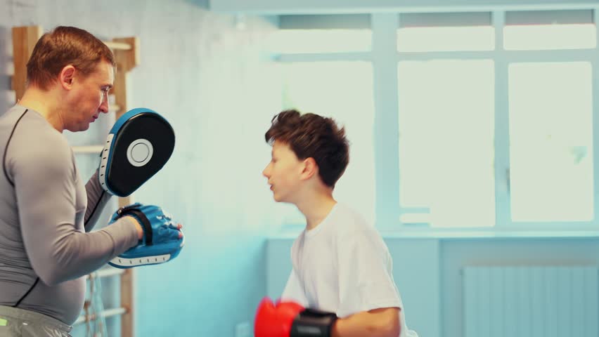 Active teen male practitioner of boxing courses applying kicks on hitting mitts during workout session