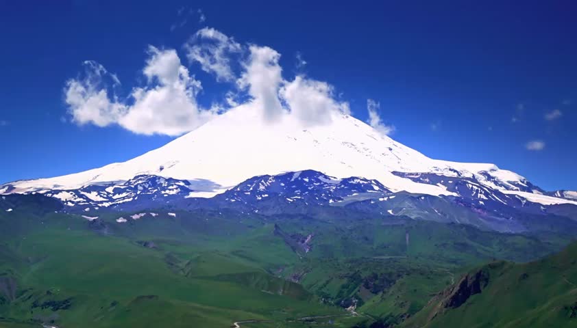 Stunning time-lapse of snow-covered mountain peak against a bright blue sky and green valley.