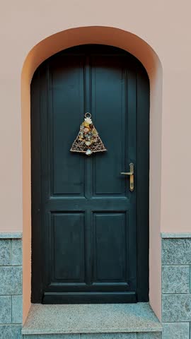 Wooden door with Christmas wreath and golden bell on Italian house facade. Festive entrance decoration for Christmas.