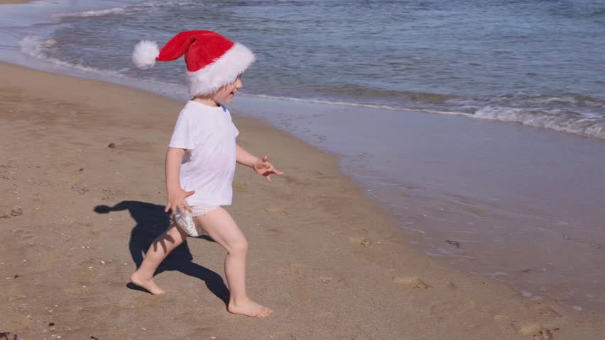 Little happy boy wearing red Santa hat running and jumping on a sandy beach near sea. Christmas holidays concept. 