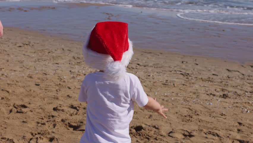 Little boy wearing red Santa hat running, jumping on a sandy beach and falling down near sea.