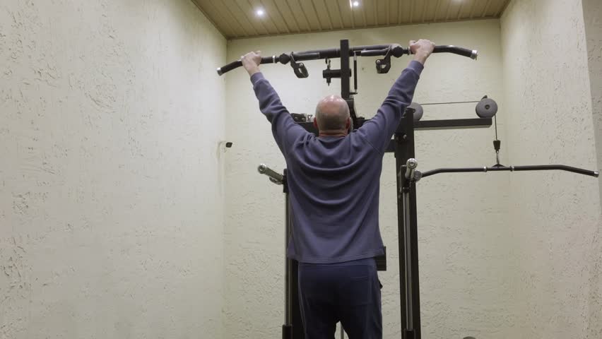 Athletic bald man in blue ribbed sweatshirt and navy pants hanging from pull up bar gripping handles on black assisted chin up station in white home gym with wooden ceiling.