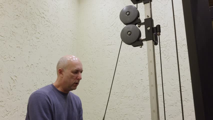 Focused bald senior man in blue sweater doing triceps cable pushdown in gym with beige textured wall background demonstrating proper form and concentration for strength training.