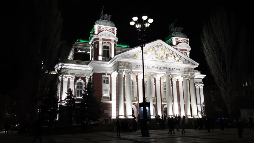 National Theatre Ivan Vazov building is brightly illuminated and stands out against evening sky. Columns and architectural elements adorn buildings facade.
