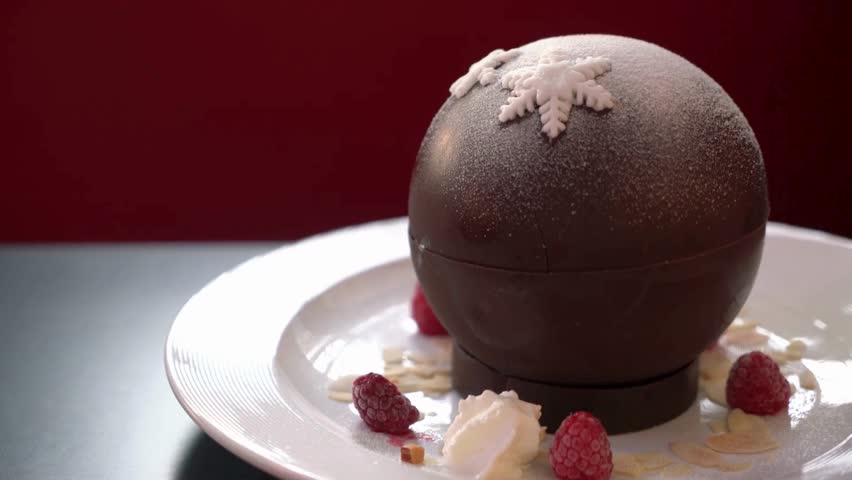Elegant chocolate sphere dessert served on a white plate with warm sauce poured over, revealing a gourmet interior. Fine dining dessert presentation.