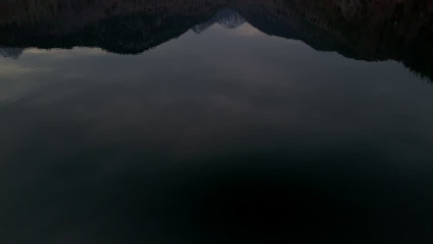 Cinematic upward-looking drone shot flying over Alpsee Lake in Füssen, Germany, during a late winter evening, with snow-capped Alpine peaks and dense forested hills surrounding the scene.