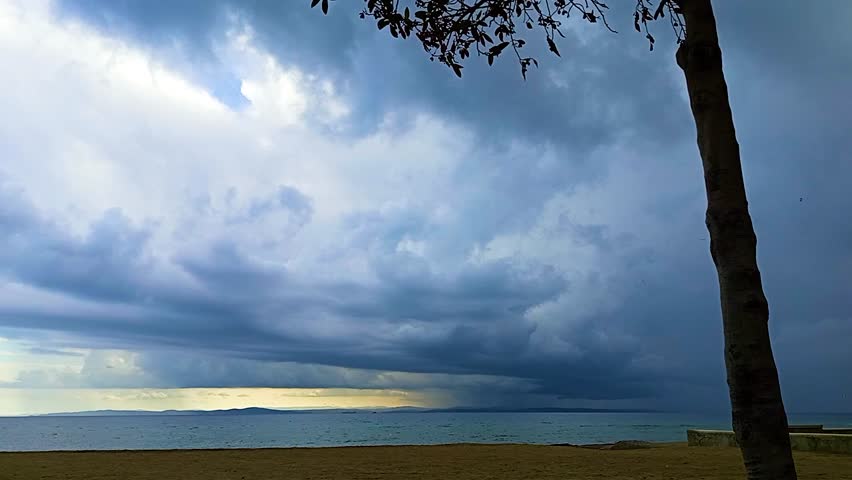 This atmospheric high-definition video captures a powerful and moody coastal scene at the Sarkoy beach in Turkey. The footage features a dramatic expanse of heavy, dark gray storm clouds rolling over the sea, creating a sense of anticipation and the raw power of nature. In the foreground, a solitary tree trunk stands tall, adding a sense of scale and grounding the composition, while its branches frame the top of the view. The horizon shows a sliver of light where the sun attempts to break through the overcast sky, casting a pale glow over the distant mountains and the calm but dark waters of the Marmara Sea.

This clip is an excellent choice for filmmakers, weather enthusiasts, and digital creators looking for high-quality "moody" or "dramatic" nature b-roll. It is perfectly suited for documentaries about climate, environmental storytelling, or as a background for contemplative and emotional cinematic projects. The contrast between the sandy shore, the dark sea, and the imposing cloud formations provides a visually striking aesthetic that symbolizes change, tranquility before a storm, or the immense scale of the natural world. Whether used for a travel vlog highlighting the diverse weather of the Thrace region or a professional marketing campaign needing an atmospheric outdoor setting, this footage offers a professional and evocative look at the Turkish coastline under a stormy sky.