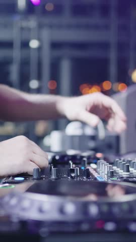 Close up of DJ hands adjusting mixer controls and performing live set on professional turntables with colorful club lights and blurred nightlife background