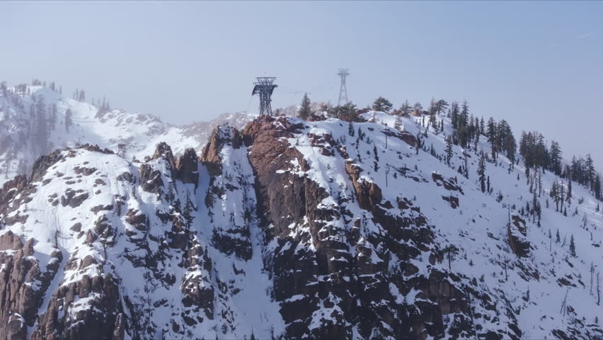 Aerial view of light snowfall over rocky ridges at Palisades Tahoe, California, USA. Snow covered cliffs, alpine terrain and scattered pine trees create a moody winter mountain atmosphere.