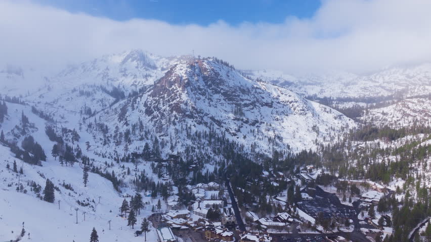 Aerial view of light snowfall over a snowy mountain resort at Palisades Tahoe, California, USA. Rocky peaks, ski slopes, pine forest and village buildings form a calm winter alpine landscape.