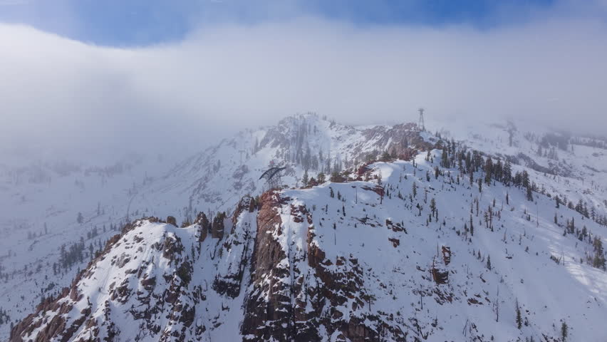 Aerial view of light snowfall drifting around a cable car tower on a rocky ridge at Palisades Tahoe, California, USA. Snow covered granite rocks and pine trees shape a rugged winter mountain scene.