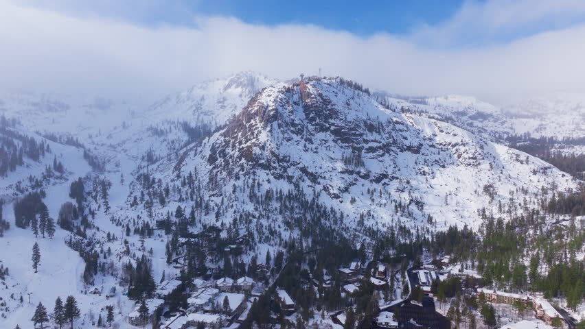 Aerial view of light snowfall over a mountain village at Palisades Tahoe, California, USA. Snow covered rooftops, ski slopes, pine forest and rocky peaks create a peaceful winter alpine scene.