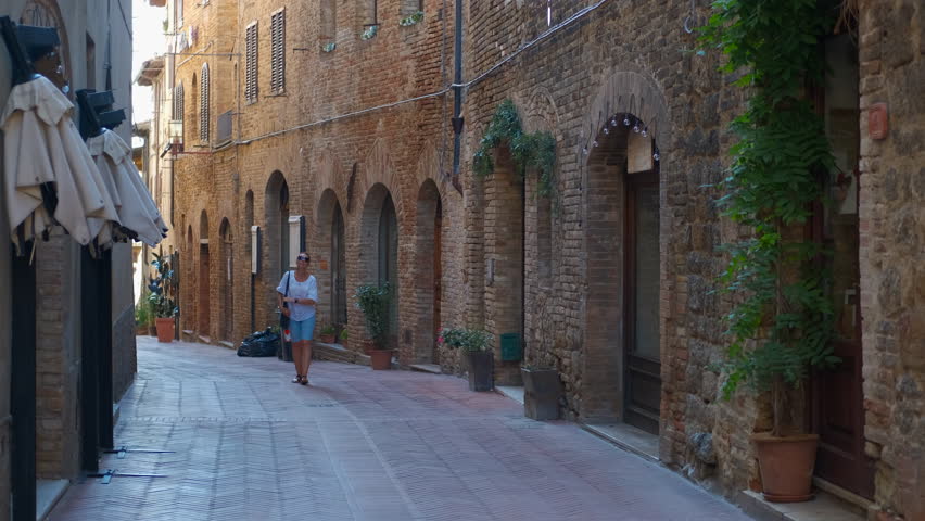 Tourist walking downhill in a medieval tuscan street. Female tourist walking downhill in a characteristic medieval tuscan street, with stone buildings and closed shops, carrying a black shoulder bag