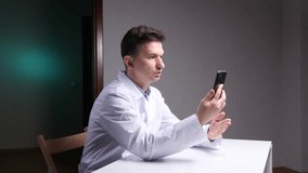 Doctor in a medical gown sits at his desk, having a virtual video consultation with a patient on his smartphone, providing online healthcare and professional medical advice - Powered by Shutterstock - Get 15% off with code: PIKWIZARD15