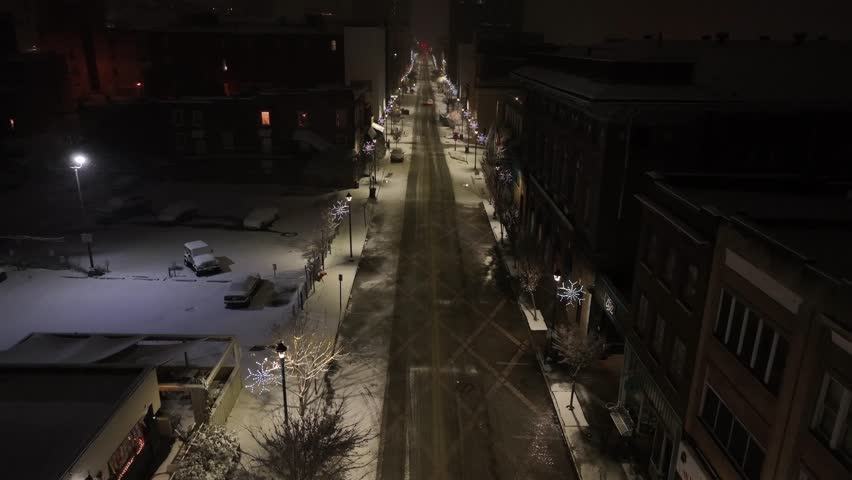 Slow flight over decorated Main Street of american town at night. Lighting snowflakes and lantern long road at soft falling snow flurries. Aerial birds eye shot.