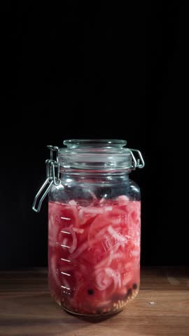 Vertical video establishing shot of hand opening a glass jar of pink pickled onions on a wooden table. Fermented food concept. Black background with copy space for text.