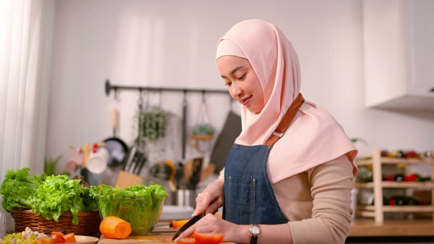 Asian Muslim woman cooking healthy foods in kitchen in morning at home. Attractive female wearing hijab, feeling happy and relax, enjoy preparing ingredients making fresh salad for breakfast in house.