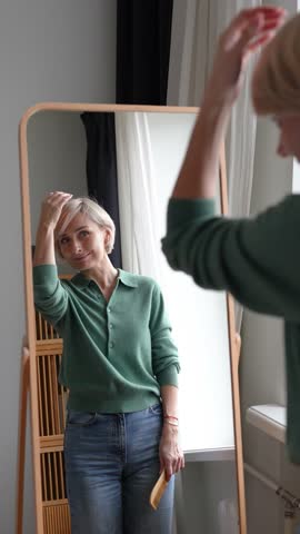 Delightful relaxed middle aged woman enjoying new haircut and hair color, standing in front of mirror in room combing hair with comb, fixing hairstyle, looking satisfied and smiling to reflection.