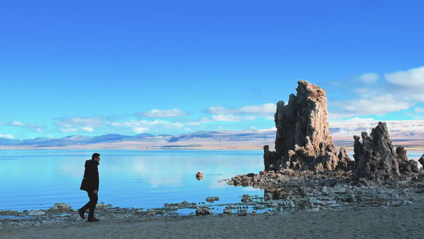 A lone traveler walks along Mono Lake’s reflective shore beside towering tufa pinnacles, with clear blue sky and distant Eastern Sierra mountains framing the otherworldly shoreline.