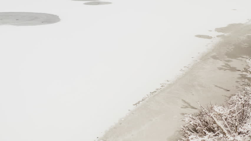 Camera tilts up from a partially frozen mountain lake to a dense pine forest and rocky hillside blanketed in fresh snow near Twin Falls, Mammoth Lakes, California.