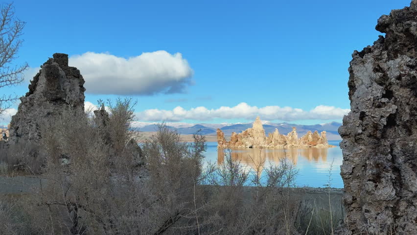 Tufa towers at Mono Lake frame a distant cluster of golden pinnacles reflected in still water, with bare desert shrubs and mountains under a clear blue sky.