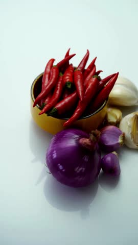Fresh red hot chilies in yellow bowl surrounded by whole and sliced red onions and garlic on white background studio shot cooking ingredients