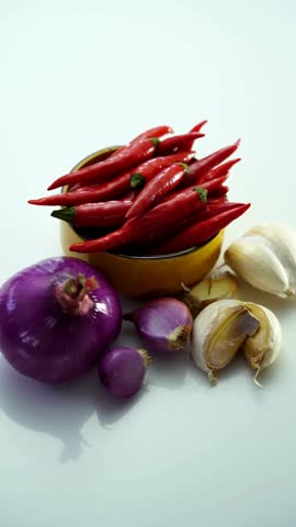 Group of fresh cooking ingredients including red chilies in yellow bowl purple onion and garlic cloves on white surface vertical shot