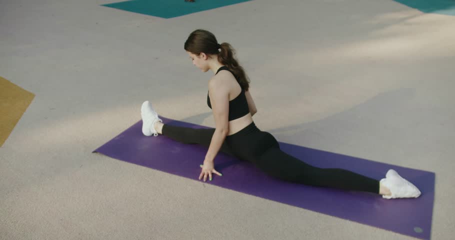 Sporty attractive female in black sportswear exercising and stretching on yoga mat outdoors. Fit girl have workout and stretch body on exercise mat. Calm woman stretching legs while practicing yoga.