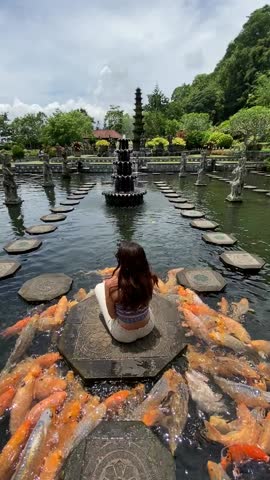 Immersive vertical video captures a tranquil scene of a woman sitting on stepping stones surrounded by colorful koi in Tirta Gangga Water Garden, Bali. Ideal for mobile and social media.