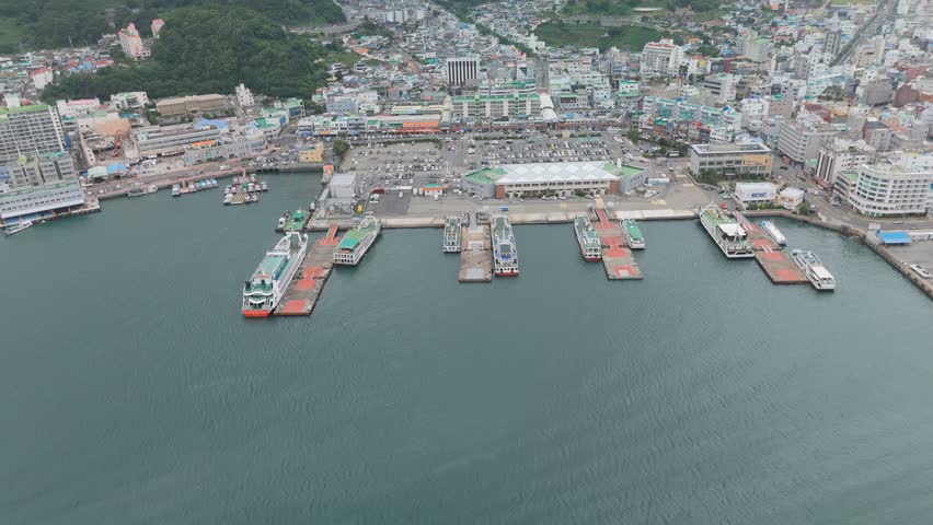 Aerial view of a bustling coastal city harbor with multiple ferries docked, surrounded by urban buildings and green hills.
