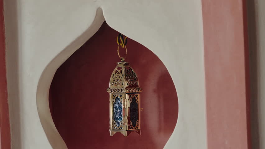 Ornate gold lantern hangs in a Moorish-style alcove. The lantern swings gently, creating a peaceful and exotic ambiance. The red clay wall and white trim add to the Moroccan feel. culture