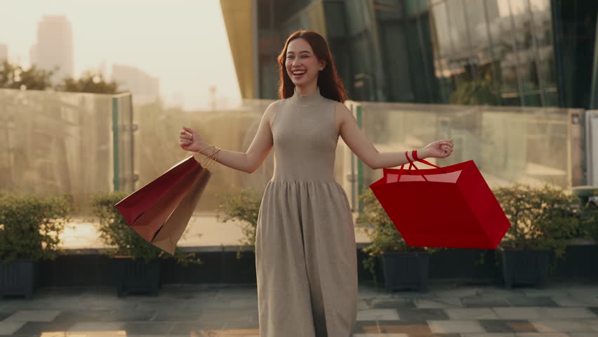 Portrait of young Asian woman dances with shopping bags bunches on business center terrace. Pretty Chinese female spins around holding store packages from sale in modern downtown