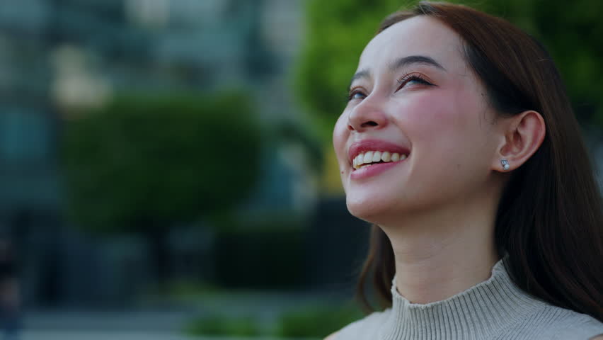 Portrait of joyful Asian woman with long loose hair smiles looking upward in park. Young Japanese lady smiles happily expressing good mood in urban garden. Positive life moments