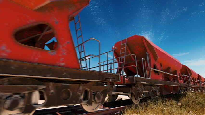 A line of weathered red hopper cars on a railway track, seen from a low angle against a clear blue sky with wispy clouds