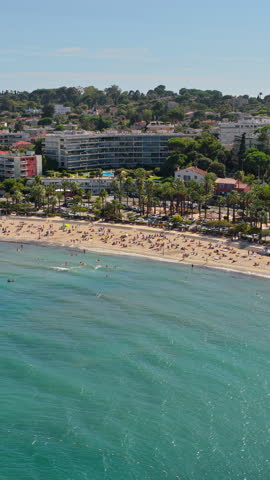 Aerial drone view of Antibes beach and promenade, lined with palm trees and apartments, with turquoise Mediterranean waters and swimmers in the bay. Vertical