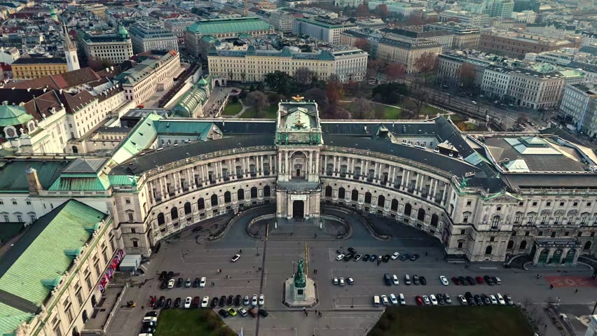 Aerial landscape drone shot panning left over Hofburg Palace in Vienna, showing the palace, surrounding cityscape, winter sunlight, cloudy skies, and tourists walking in the palace grounds below.