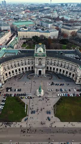 Aerial portrait drone shot panning left over Hofburg Palace in Vienna, showing the palace, surrounding cityscape, winter sunlight, cloudy skies, and tourists walking in the palace grounds below.