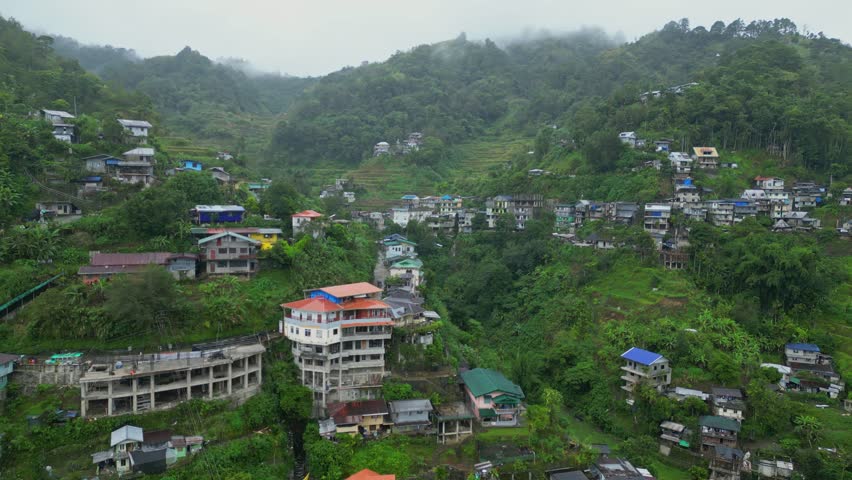 Wide pan aerial of the sprawling Banaue landscape, showcasing dense mountain ranges, tiered rice terraces, and rural villages under a misty Ifugao, Philippine sky.