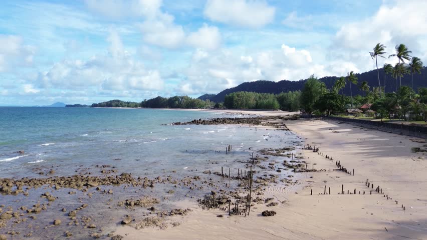 Aerial drone footage of a rocky shoreline at Long Beach Koh Lanta, Thailand, featuring lush palm trees in the foreground and tropical bungalows along the coast, with turquoise waters on a cloudy day.