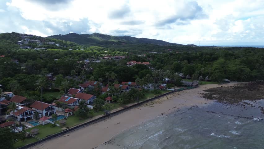 Aerial drone footage of a rocky shoreline at Long Beach Koh Lanta, Thailand, featuring lush palm trees in the foreground and tropical bungalows along the coast, with turquoise waters on a cloudy day.