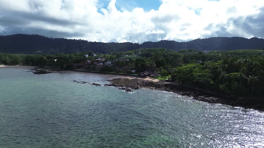 Aerial drone footage of a rocky shoreline at Long Beach Koh Lanta, Thailand, featuring lush palm trees in the foreground and tropical bungalows along the coast, with turquoise waters on a cloudy day.