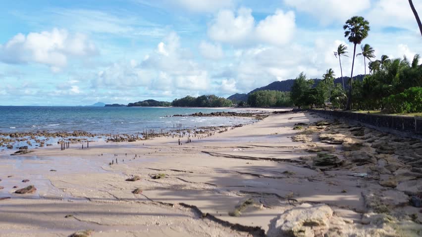 Aerial drone footage of a rocky shoreline at Long Beach Koh Lanta, Thailand, featuring lush palm trees in the foreground and tropical bungalows along the coast, with turquoise waters on a cloudy day.