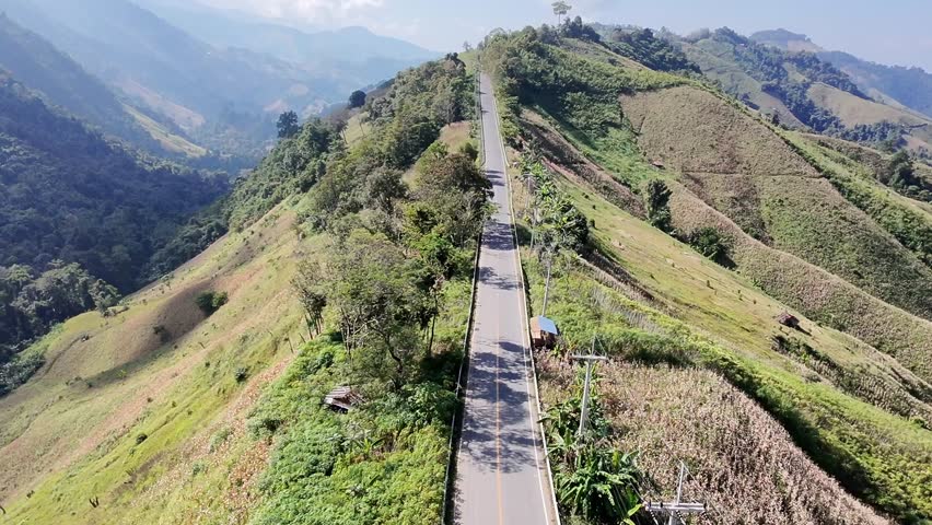 Aerial drone view of a winding mountain road along a green ridge, surrounded by rolling hills, forests, and rural farmland. Scenic landscape showing travel, freedom, infrastructure, and nature