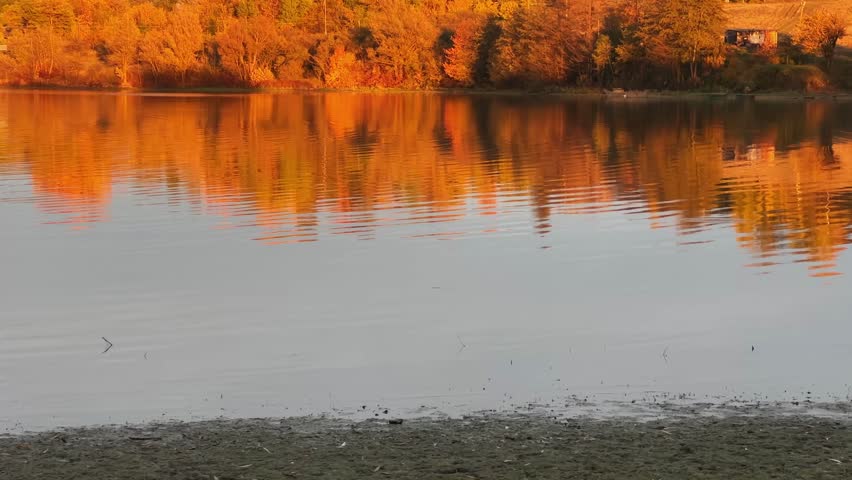 Pond with calm water surface in valley with hilly opposite bank partially overgrown with trees against the cloudy sky, view while vertical panning in sunny evening during sunset
