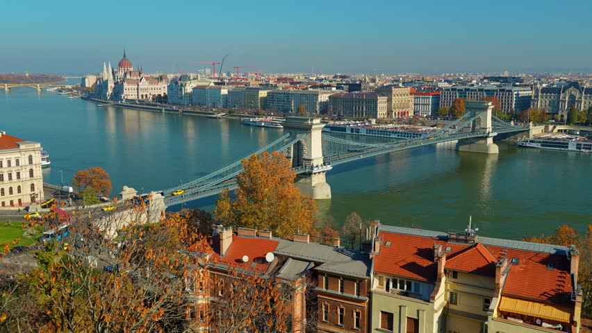 Aerial view of the historic Szechenyi Chain Bridge crossing the Danube river in Budapest, Hungary, surrounded by colorful autumn trees and classic European architecture
