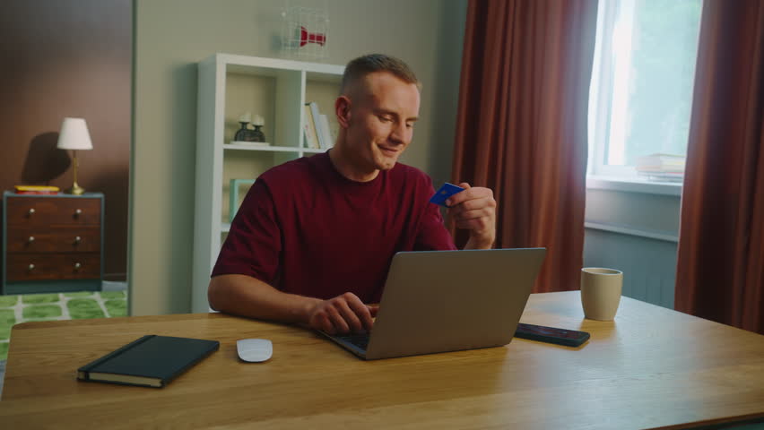 Young adult man sit desk home pay by bank credit card buy stuff online use laptop computer, mobile cell phone. Smiling person do purchase internet store. Remote shopping. Big holiday sale, discounts.