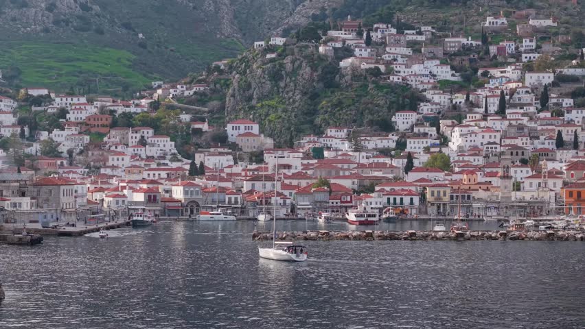 Drone ascends from the sea to reveal Hydra Port as a sailing boat approaches, capturing the island harbor and its coastal landscape.