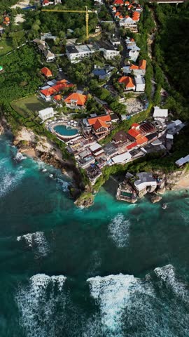 Vertical aerial footage capturing dramatic Uluwatu cliffside restaurants glowing in golden hour light above calm blue ocean creating a peaceful tropical sunset atmosphere.