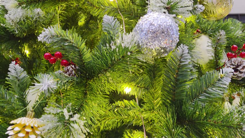 Slow motion close-up of a decorated Christmas tree with warm fairy lights, frosted pine branches, red berries, and silver ornament. Cozy festive holiday background in high definition