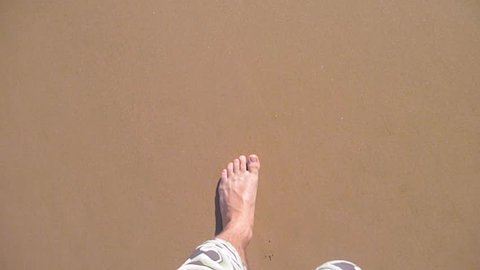 Man walking bare foot on sandy beach into ocean wave, point of view.